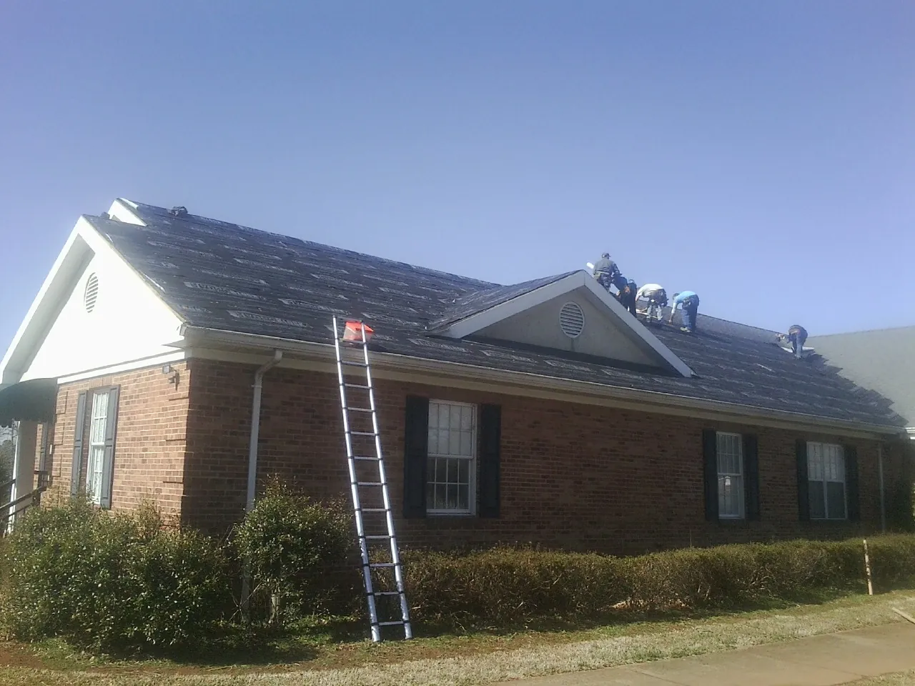 Crew tearing shingles off a brick ranch roof with a ladder set — Emergency Roof Repair work by our crew in Magee