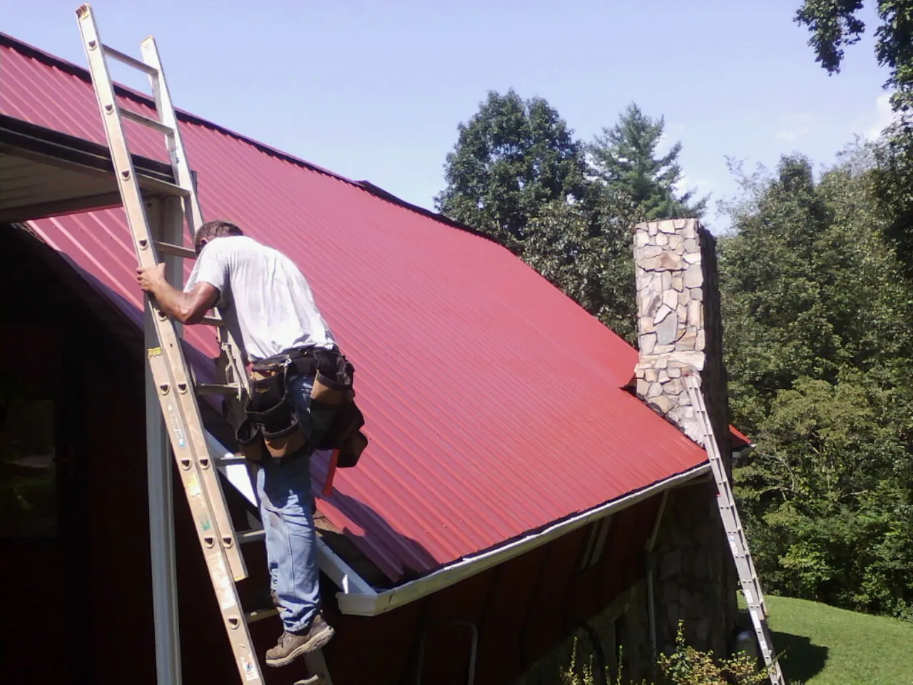 Roofer on a ladder beside a red metal roof and stone chimney — Soffit & Fascia Repair by Roofing Brothers in Magee